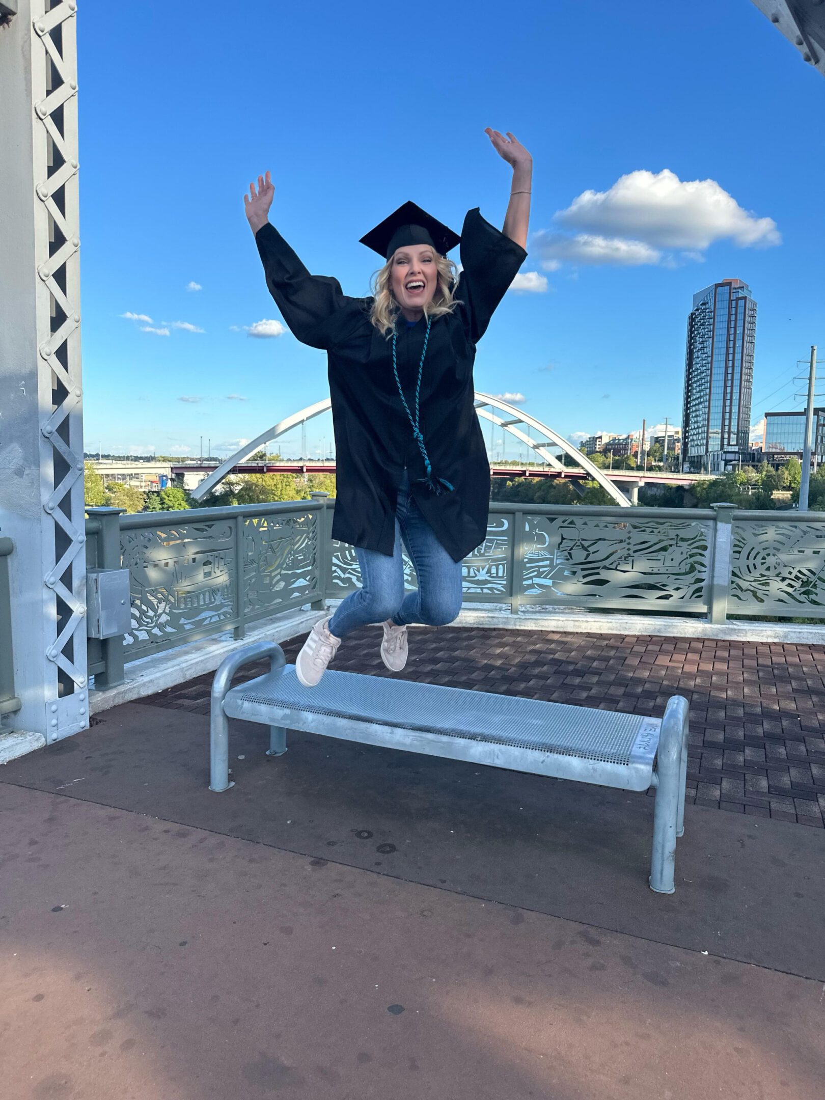 Graduate in cap and gown jumping joyfully on a bench outdoors.